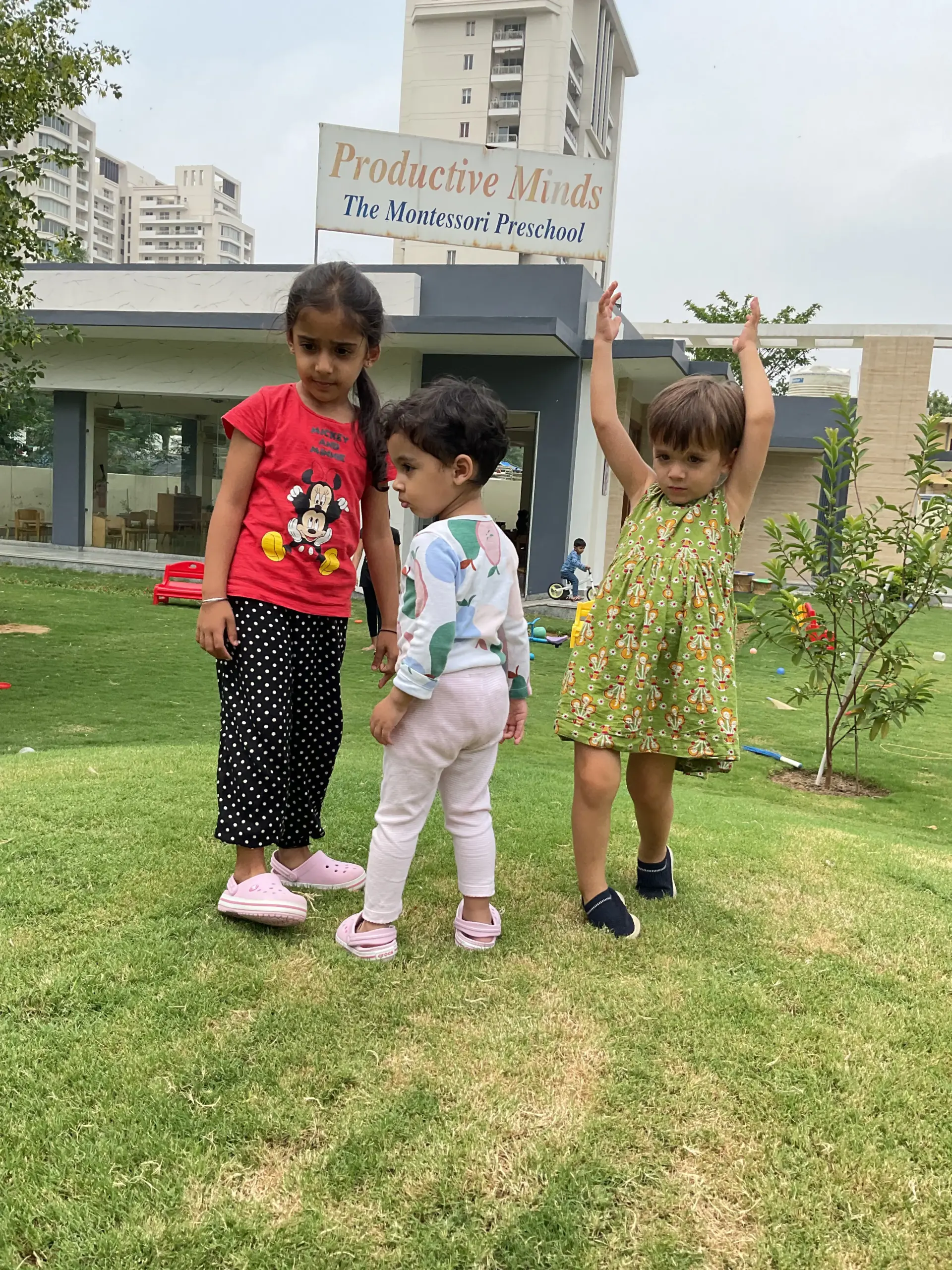 Three children playing on green grass in front of preschool building