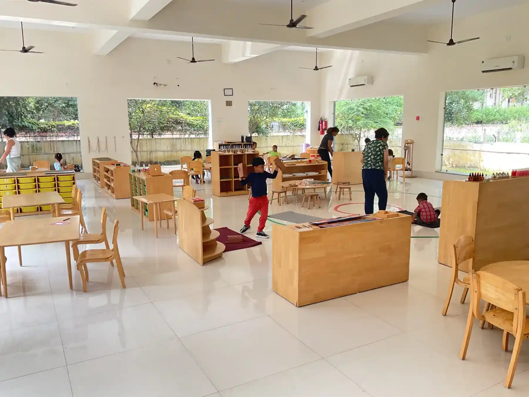 Children working peacefully at wooden tables in bright sunlit Montessori classroom with floor-to-ceiling glass windows