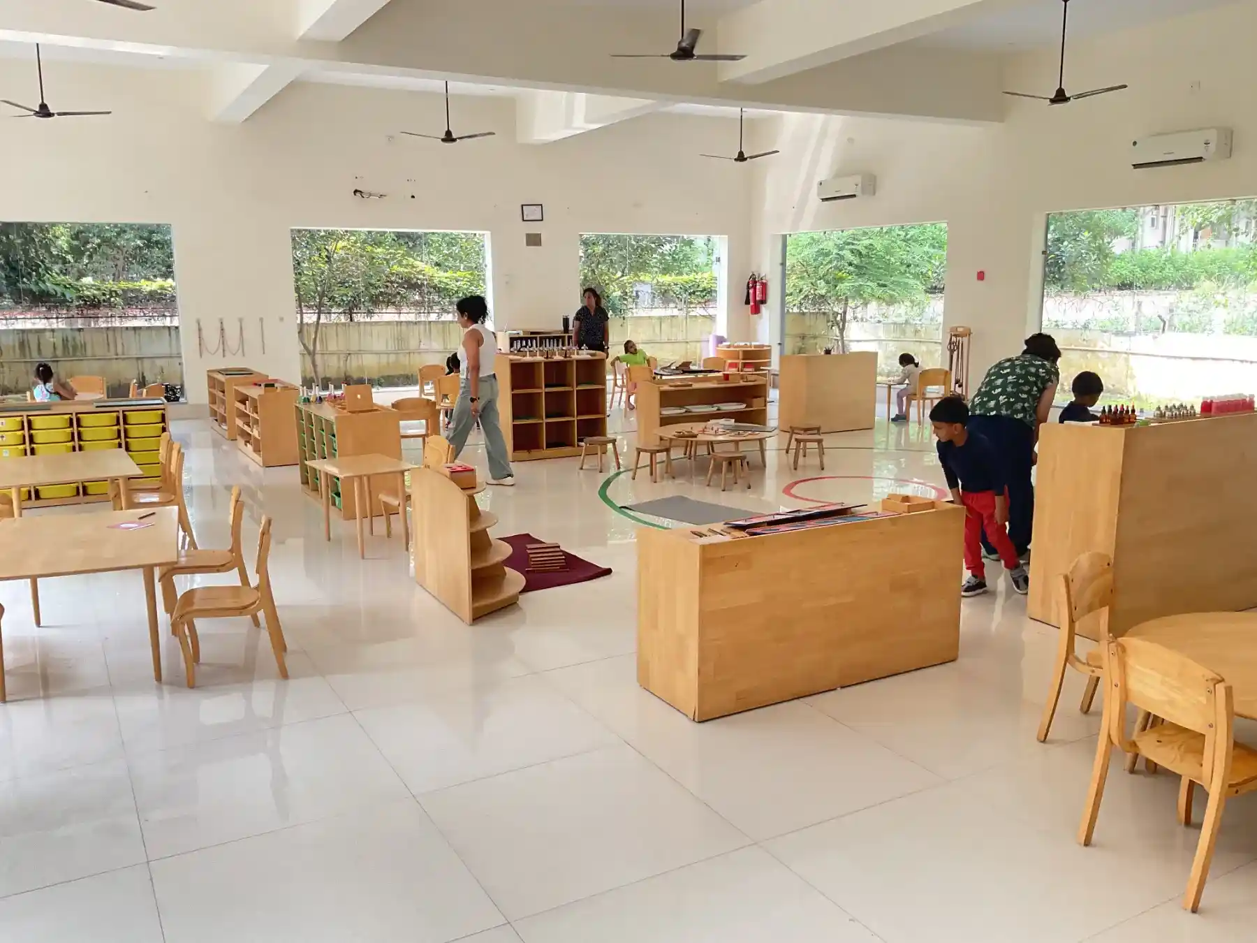 A bright, sunlit Montessori classroom with floor-to-ceiling glass windows and children working at wooden tables