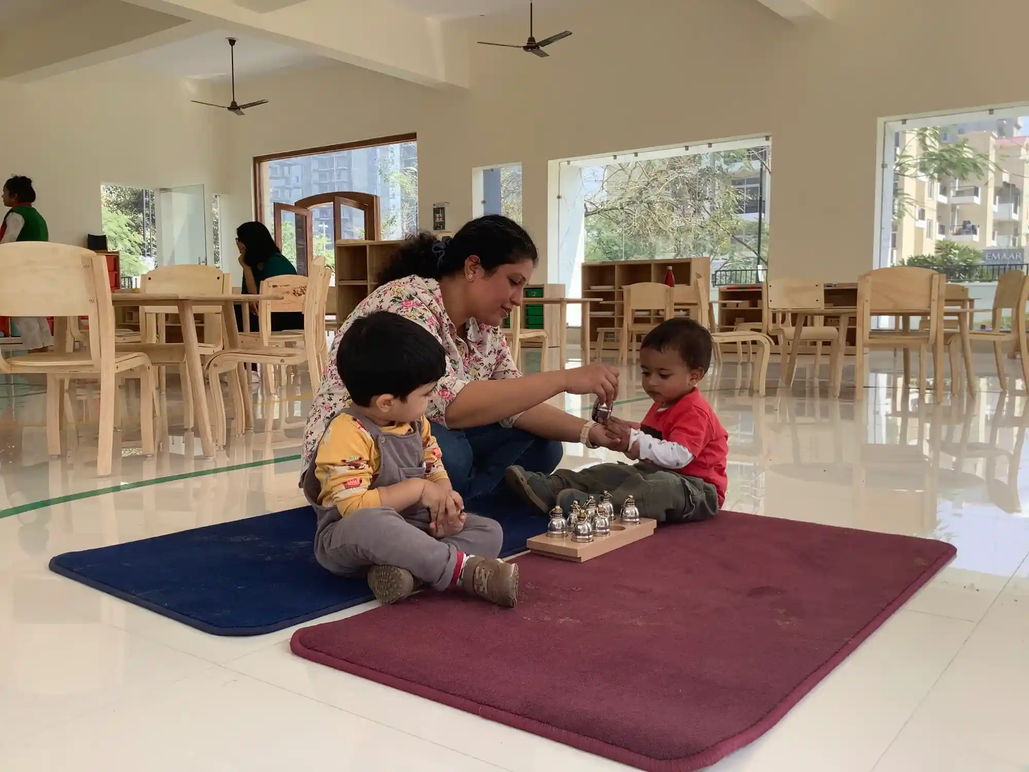 A bright, sunlit Montessori classroom with floor-to-ceiling glass windows and children working at wooden tables